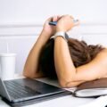 A woman resting her head on her desk beside a laptop, overwhelmed
