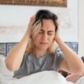 A woman sitting up in bed holding her head after waking