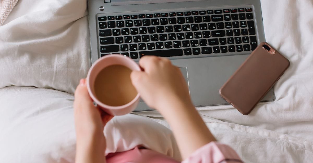 Hands holding a coffee mug beside a laptop on a cozy bed